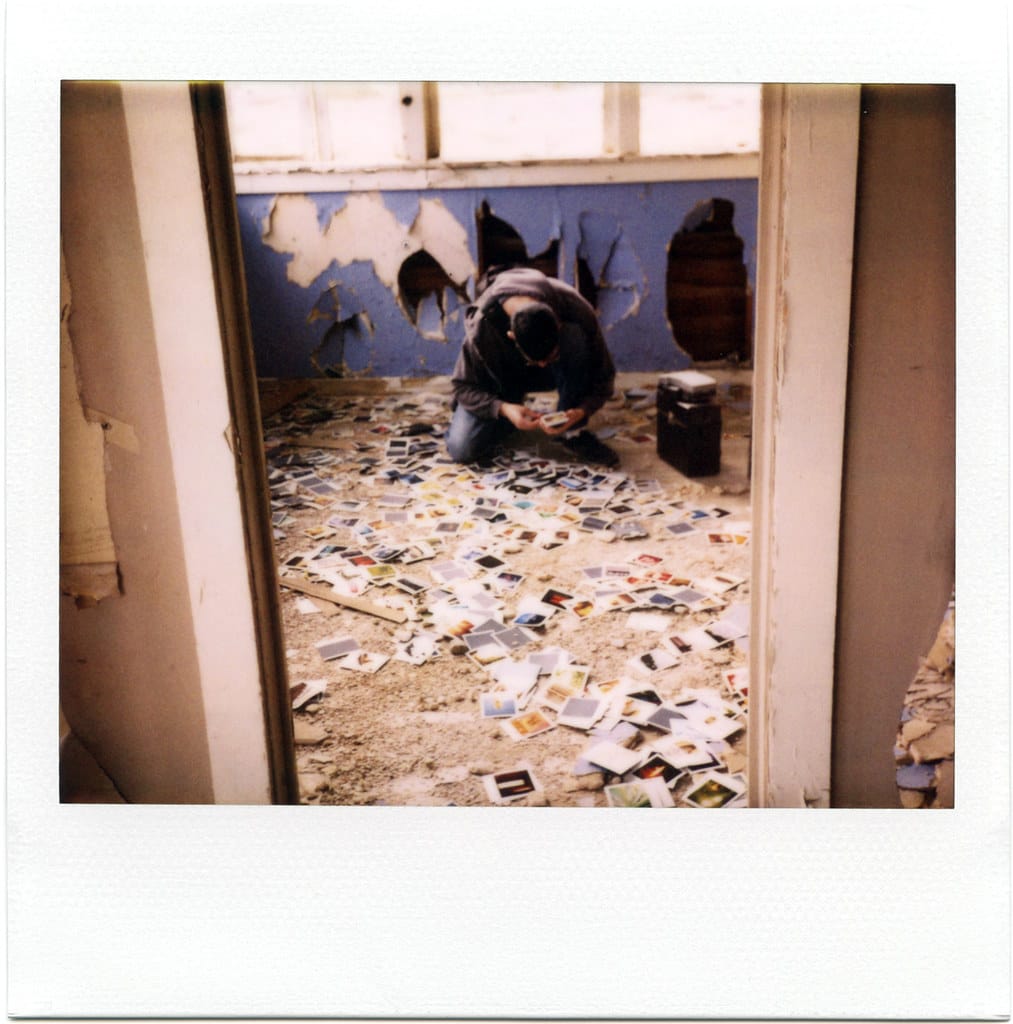 a polaroid of a man kneeling in an abandoned house surrounded by hundreds of polaroid pictures