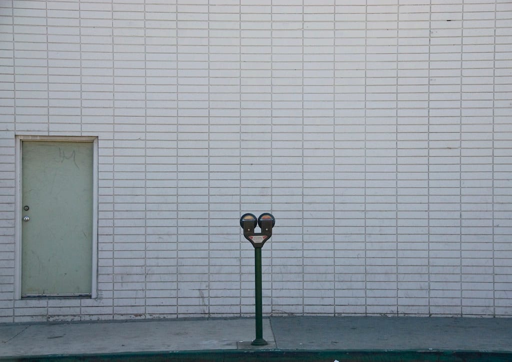 a photo of a double-headed parking meter in front of a wall made of rectangular white bricks with a single pale green door 