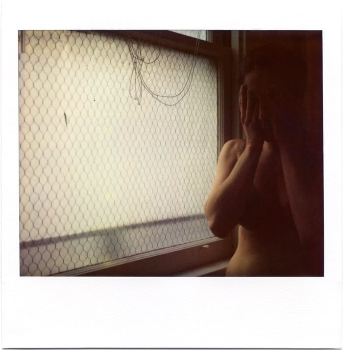 a polaroid of a woman with a bare torso covering her face with her hands next to a window